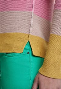 Maglione in maglia con righe rosa, beige e gialle; presenta un orlo spaccato. Abbinato a pantaloni verdi, con dettaglio di tessuto strutturato.