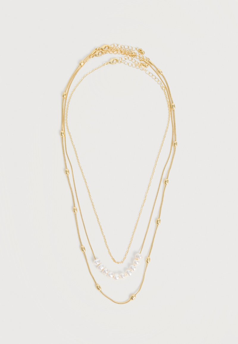 Τριπλό κολιέ σε χρυσό τόνο με μικρές χρυσές χάντρες σε δύο αλυσίδες και λευκές πέρλες στην κοντύτερη αλυσίδα, παρουσιασμένο σε λευκό φόντο.