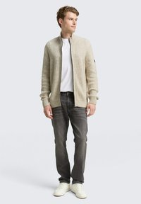 Beige gebreide cardigan met een geribde kraag, ritssluiting en lange mouwen, gecombineerd met een wit shirt, donkere jeans en witte sneakers.