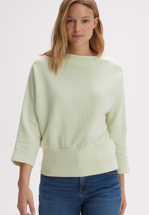 Light green ribbed pullover with a boat neckline and three-quarter sleeves. Soft texture and a fitted waistband. Paired with dark jeans.