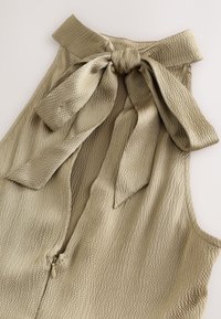 Beige gestructureerde mouwloze top met een hoge kraag die in een grote strik is gebonden en een ritssluiting aan de voorkant.