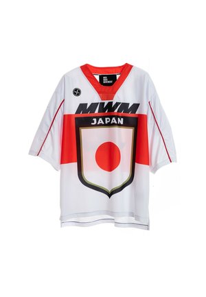 Joven con pelo trenzado que lleva una camiseta blanca y roja con un diseño de Japón, pantalones cortos negros, calcetines negros y botas marrones, de pie sobre un fondo blanco.