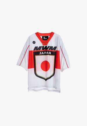 Joven con pelo trenzado que lleva una camiseta blanca y roja con un diseño de Japón, pantalones cortos negros, calcetines negros y botas marrones, de pie sobre un fondo blanco.
