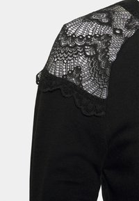 Černý svetr se strukturovaným krajkovým ramenním panelem se zvlněným okrajem a složitými květinovými vzory. Měkká pletenina s hladkým povrchem.