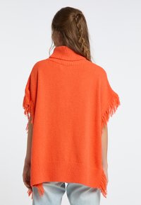 Poncho naranja de punto con un alto cuello acanalado, bordes flecos y un corte relajado. La tela parece suave y texturizada.