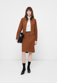 Donna con giacca in camoscio marrone e gonna abbinata, top bianco, scarpe e calze nere, che tiene una grande borsa a mano nera.