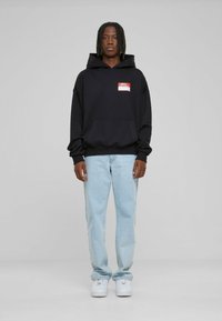 Black hooded sweatshirt with front pocket and red logo patch, paired with light blue jeans and white sneakers. Model stands facing forward.