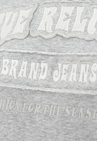Szara tkanina z haftowanym białym tekstem "LOVE RELIGION BRAND JEANS". Posiada przeszycia akcentujące oraz teksturowaną powierzchnię.