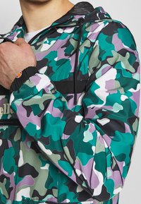 Camouflagejack met een mix van teal, paars en zwart. Gemaakt van lichtgewicht stof, met elastische manchetten en een ritssluiting aan de voorkant.