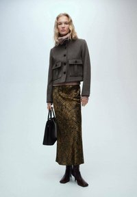 Braune Woll-Croppjacke mit zwei Fronttaschen, kombiniert mit einem langen Rock im Gold- und Schwarz-Schlangenprint. Schwarze Tasche und burgunderfarbene Absatzstiefel.