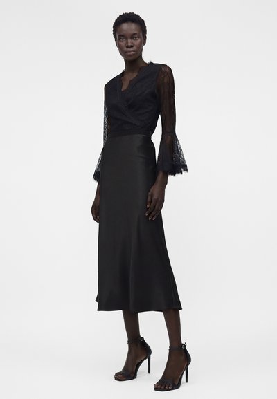 Γυναίκα που φοράει μαύρο δαντελωτό τοπ τύπου wrap με μανίκια καμπάνα και μια μαύρη σατέν φούστα midi, συνδυασμένα με μαύρα πέδιλα με ψηλό τακούνι.