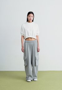 Sweatshirt cropped branco com design em relevo, combinado com calças cargo oversized cinzas com bolsos laterais, tudo sobre um chão verde.