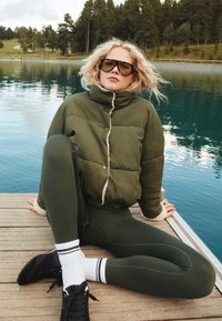 Zielona pikowana kurtka z wysokim kołnierzem i kontrastową kremową podszewką, zestawiona z ciemnozielonymi legginsami i czarnymi sneakersami z białymi akcentami.