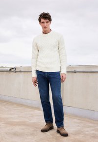 Weißer Kabelstrickpullover mit Rundhalsausschnitt, kombiniert mit blauen Jeans und braunen Schnürschuhen, stehend auf einer Betonoberfläche.