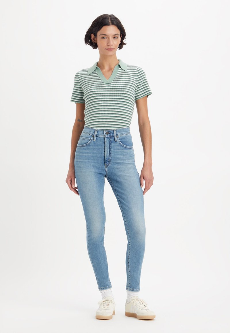 Junge Frau in einem grün gestreiften Kurzarmhemd, mit hoch taillierten hellblauen Jeans, weißen Socken und weißen Turnschuhen, stehend.