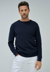 Maglione lavorato a maglia blu navy con scollo rotondo, maniche lunghe e un motivo testurizzato. Abbinato a pantaloni bianchi, enfatizzando uno stile casual.