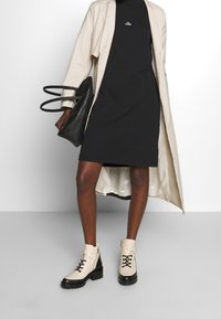 Personne portant une robe noire, un trench-coat beige, des bottes à lacets crème et noires, tenant un sac à main noir avec des poignées circulaires.