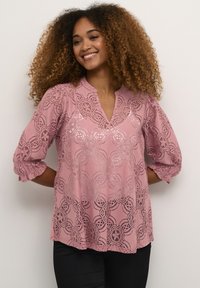 Blouse en dentelle rose avec un col en V, manches bouffantes trois-quarts, motifs floraux en découpe, et détails texturés à l'ourlet et aux manches.