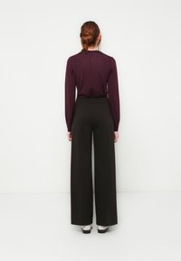 Langärmliges lila Oberteil kombiniert mit hoch taillierten, schwarzer, weit geschnittenen Hosen, die eine glatte Textur und eine klare, schlichte Silhouette zeigen.