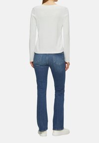 Weißes Langarmshirt mit abgerundetem Saum, kombiniert mit blauen Schlagjeans und weißen Sneakers, von hinten betrachtet.