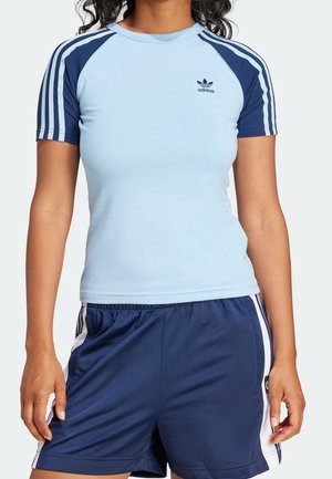Kobieta ubrana w jasnoniebieską koszulkę Adidas z granatowymi rękawami oraz pasujące granatowe krótkie spodenki sportowe z białymi paskami.