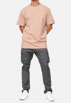 Homme portant un t-shirt à manches courtes rose clair, un pantalon cargo gris avec poches et des baskets blanches, debout les mains derrière le dos.