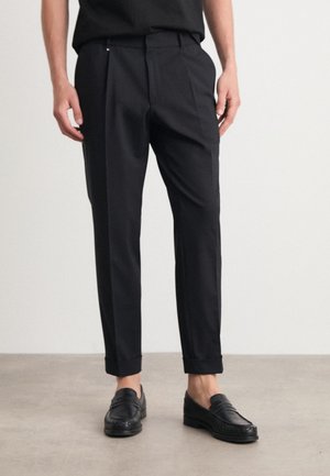 Black tailored trousers with a straight fit, smooth fabric, subtle pleats, and a rolled hem. Paired with black loafers, on a neutral background.