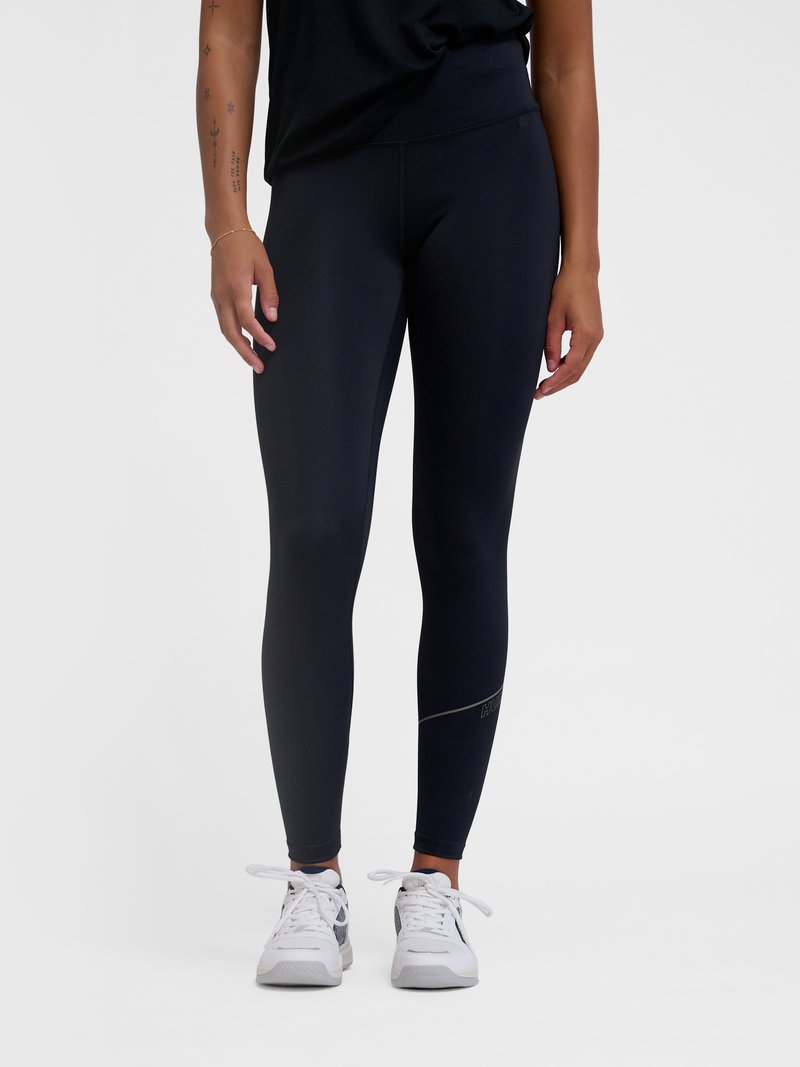 Jalad mustade kitsaste retuuside ja valgete tossudega, käed lõdvestunult külgedel, ühtlase heleda taustaga.