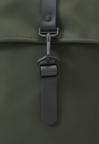 Gros plan sur un tissu imperméable vert foncé avec une sangle en caoutchouc noir et un fermoir en métal étiqueté « RAINS ». 