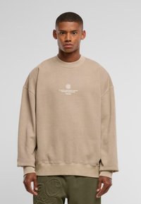 Beige sweatshirt med rund hals og løs pasform, med et hvidt trykt logo og tekst på forsiden. Blød tekstur og ribbede manchetter.
