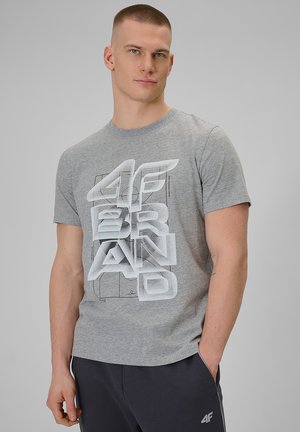Młody mężczyzna o krótkich włosach, ubrany w szary t-shirt z nadrukiem i ciemne spodnie, stoi z jedną ręką w kieszeni na jednolitym tle.