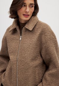 Veste marron texturée avec fermeture éclair à l'avant, grand col et coupe décontractée. Présente de subtils motifs en vagues sur le tissu et des poches latérales.