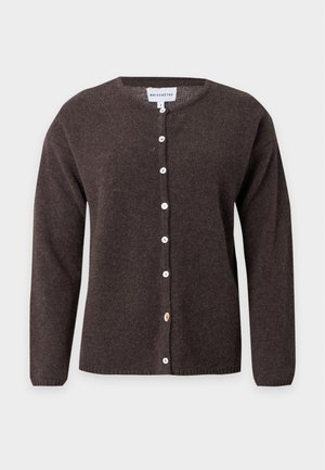 Kvinde med langt blondt hår iført en mørkebrun knappet cardigan, hvid skjorte, beige bukser med vide ben og brune sko, stående med den ene hånd på hoften.