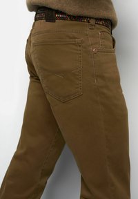 Braune Hose mit detaillierter Stickerei auf der Gesäßtasche, getragen mit einem mehrfarbigen, gewebten Gürtel und einem braunen Pullover, der am Bund sichtbar ist.