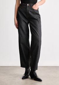 Jeans de denim cinza escuro e brilhante, com corte solto, cintura alta, cinco bolsos e costura laranja em contraste. Usados com botas pretas até o tornozelo.