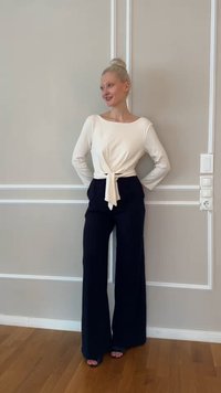 Γυναίκα με ξανθά μαλλιά σε κότσο, που φοράει λευκό μακρυμάνικο τοπ με δέσιμο μπροστά και μαύρο παντελόνι με φαρδιά γραμμή, στέκεται μπροστά σε ένα φωτεινό γκρι τοίχο με πάνελ.