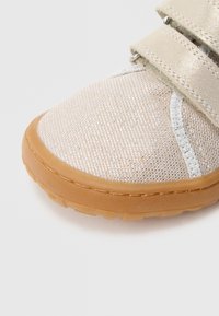 Beige athletic shoe made of mesh with a textured surface, featuring a tan rubber sole and two Velcro straps.