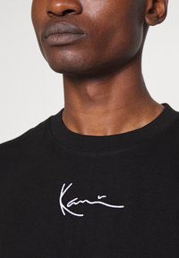 Black cotton t-shirt featuring a crew neckline and a white embroidered signature on the chest, showcasing a smooth texture.