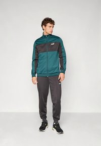 Sledenjska jakna v barvi zeleno-modre in črne, iz poliestra, z zadrgo spredaj in logotipom. Skupaj s temnimi hlačami in črnimi športnimi copati.