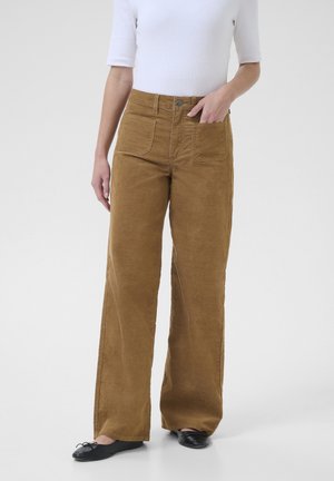 Kobieta ubrana w brązowe sztruksowe spodnie z wysokim stanem i szerokimi nogawkami oraz czarne baleriny, z jedną ręką w przedniej kieszeni, stojąca na białym tle.