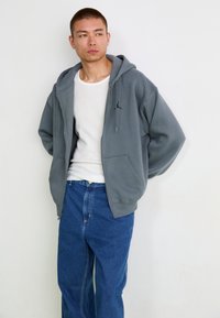 Gray zip-up hoodie with a kangaroo pocket and drawstring hood, featuring a black logo on the left chest. Paired with blue jeans.