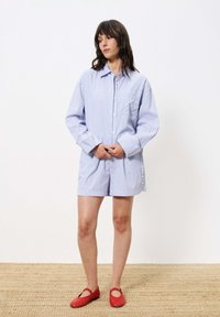 Gestreiftes blaues Hemd mit Knöpfen und einer Tasche, kombiniert mit passenden Shorts und roten Schuhen. Das Outfit zeichnet sich durch einen lockeren Schnitt und ein klassisches Design aus.