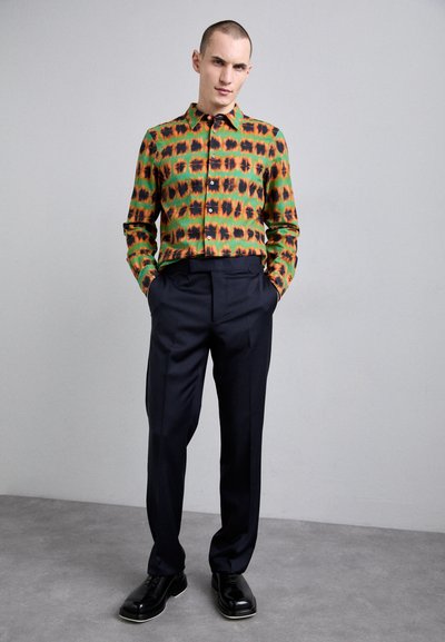 Chemise multicolore avec des motifs verts, oranges et noirs, assortie à un pantalon noir bien taillé et des chaussures noires. Texture de tissu lisse.