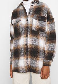 Hnědá, černá a bílá károvaná košile s knoflíky a dvěma náprsními kapsami, nošená přes bílý top a spárovaná s světle béžovými kalhotami.