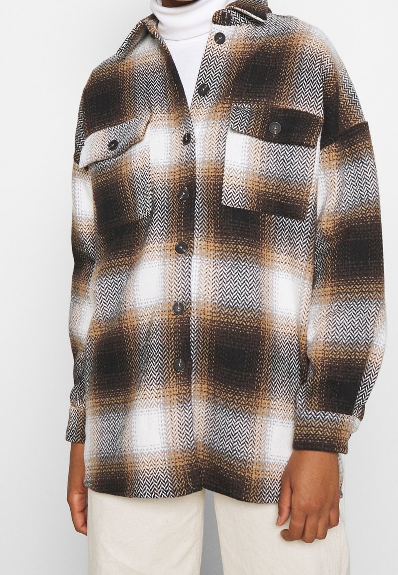 Hnědá, černá a bílá károvaná košile s knoflíky a dvěma náprsními kapsami, nošená přes bílý top a spárovaná s světle béžovými kalhotami.