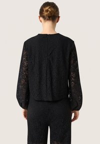 Top in pizzo nero con maniche lunghe a sbuffo, dotato di scollatura rotonda e dettaglio con zip sul retro. Motivo in pizzo floreale testurizzato su tutta la superficie.