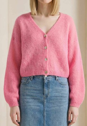 Femme portant un cardigan rose duveteux boutonné et une jupe en jean bleue taille haute, debout devant un fond neutre.