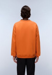 Felpa arancione con vestibilità comoda, colletto e polsini a costine, caratterizzata da una texture liscia e un design semplice. I pantaloni neri completano l'aspetto.