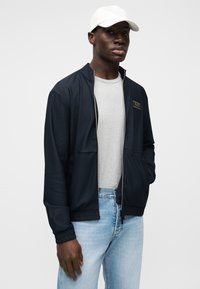 Μπουφάν τύπου track με φερμουάρ σε ναυτικό μπλε χρώμα, με υφή, μαύρες λεπτομέρειες και χρυσή επιγραφή λογότυπου, συνδυασμένο με γκρι μπλούζα και ανοιχτό μπλε τζιν.