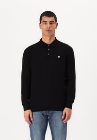 Svart långärmad polo med texturerat tyg, tre-knappskn�äppning och en gul örnlogotyp på vänster bröst. Bärs med blå jeans.